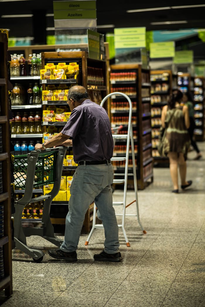 An elderly man shopping with a cart in a grocery store aisle filled with various products.